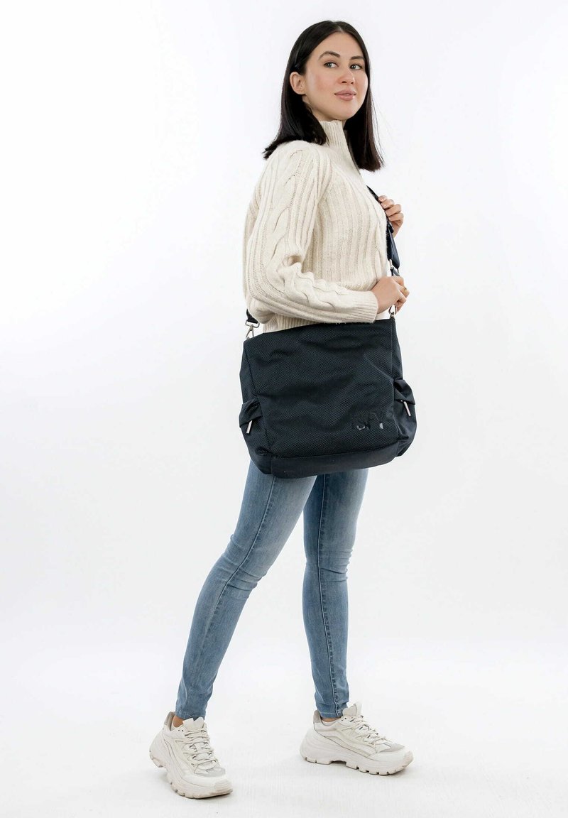 Borsa a spalla in tessuto nero con superficie texturizzata, dotata di tracolla regolabile e tasche con zip, indossata sulla spalla di un modello.