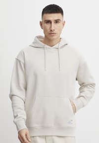 Svetlo béžová mikina vyrobená z mäkkého materiálu, ktorá má klokanie vrecko, šnúrky na kapucni a rebrované manžety. Minimalistický dizajn, voľný strih.