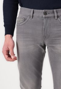 Graue Jeans aus Denim mit leichtem Stretch, regulärer Passform, Fünf-Taschen-Design und einem Logo-Patch auf der Gesäßtasche. Die Textur wirkt glatt.