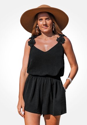 Czarny kombinezon bez rękawów wykonany z teksturowanej tkaniny, z kwiatowymi zdobieniami na ramiączkach i elastycznym paskiem w talii. Noszony z kapeluszem o szerokim rondzie.