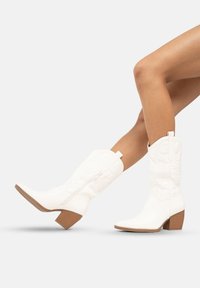 Botas de vaquero blancas con un acabado texturizado, punta afilada, tacón bloque de altura media y detalle de cremallera lateral. Acentuadas con una tira para tirar.