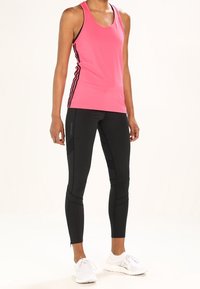 Różowy top na ramiączkach z czarnymi paskami po bokach, zestawiony z czarnymi legginsami z teksturowanymi panelami i zamkami przy kostkach, noszony z białymi butami sportowymi.