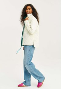 Biela prešívaná bunda s vysokým golierom, vrstvená nad zelenou košeľou. Skombinovaná s svetlomodrými džínami a ružovými teniskami s hnedými podrážkami.