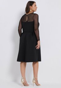 Nainen polvipituista mustasta mekosta, jossa on läpinäkyvät pitkät hihat ja selässä aukko, kultaisten korkokenkien ja kukka-korvasten kanssa.