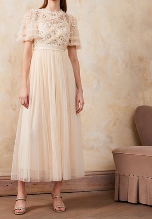 Frau trägt ein hellbeiges Tüllkleid mit Blumenstickerei auf den Puffärmeln und einem plissierten Rock, steht neben einem beigen Stuhl.