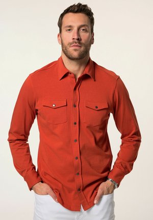 Rotes Langarm-Hemd mit Knöpfen, zwei Brusttaschen und schwarzen Druckknöpfen. Uni-Farbe mit strukturiertem Stoff. Modell trägt weiße Hosen.
