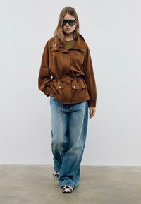 Veste en daim marron avec fermeture éclair à l'avant, taille adjustable à cordon, et deux poches avant, associée à un jean léger à jambes larges bleu clair et des chaussures à motifs.
