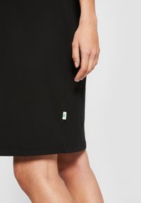 Czarna sukienka z gładkiego materiału, o długości do kolan, z małą białą metką z zielonym liściem u dołu po lewej stronie.