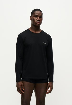 Junger Mann, der ein schwarzes Langarmshirt mit dem Wort "BOSS" auf der Brust und schwarze Shorts trägt, steht vor einem schlichten hellen Hintergrund.