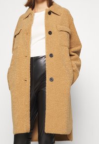 Tan faux shearling coat with a button-front design, two chest pockets, and a relaxed fit, paired with black leather pants.