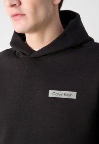 Czarna bawełniana bluza z kapturem z dużą kieszenią i szarym prostokątnym metkiem z napisem "Calvin Klein" w kolorze czarnym. Miękka tekstura i regulowany kaptur.