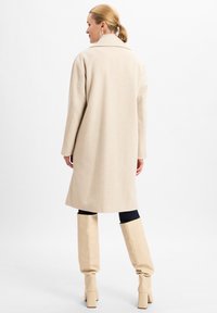 Beige visgraatjas met een brede kraag, lange mouwen en een rechte snit, gecombineerd met hoge crèmekleurige laarzen en donkere leggings.