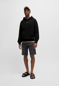 Zwarte hoodie met voorzak, grijze denimshorts en zwarte sandalen. Model draagt een lichtgrijze pet. Eenvoudig, casual ontwerp.