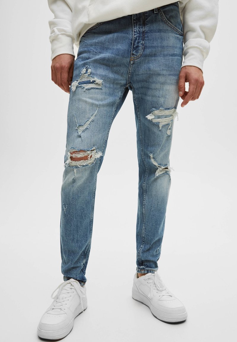 Man med slitna blå jeans och vita sneakers mot en enfärgad vit bakgrund, med avslappnade händer längs sidorna.