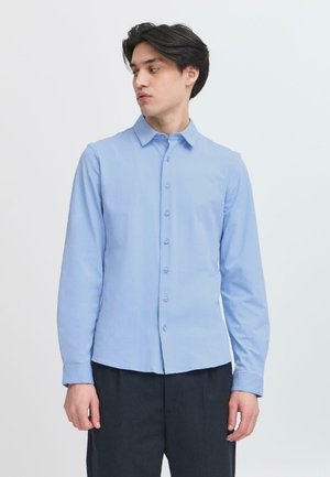 Camisa de manga larga azul claro con cierre de botones en la parte frontal, cuello puntiagudo, textura suave y corte entallado. Combinada con pantalones oscuros.