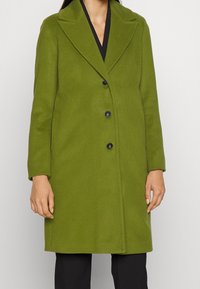 Grüner Wollmantel mit Reverskragen, drei Knöpfen und langen Ärmeln. Glatte Textur, knielang, mit tailliertem Schnitt.