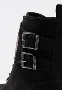Stivale ankle in pelle scamosciata nera con due fibbie metalliche, superficie testurizzata e design robusto. Nota trama a scacchi sul lato.