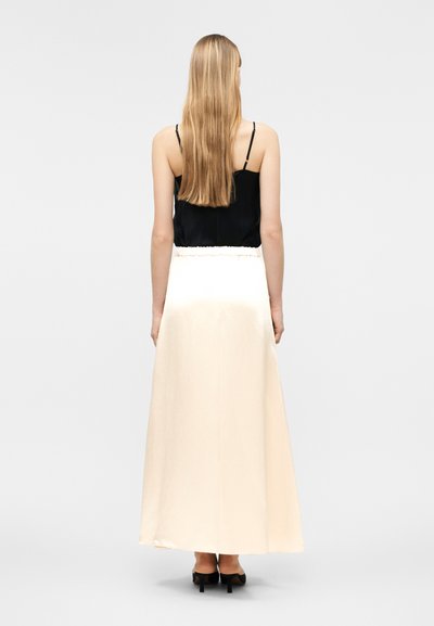 Γυναίκα με μαλλιά μακριά ξανθά, που φοράει μαύρο αμάνικο τοπ, κρέμ μακριά φούστα και μαύρα ψηλοτάκουνα, στέκεται γυρισμένη πλάτη σε απλό φόντο.