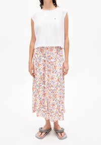 Kvinna som bär en ärmlös vit crop top, en blommig midikjol med röda, lila och gröna blommor, samt grå skum-sandaler, stående mot vit bakgrund.