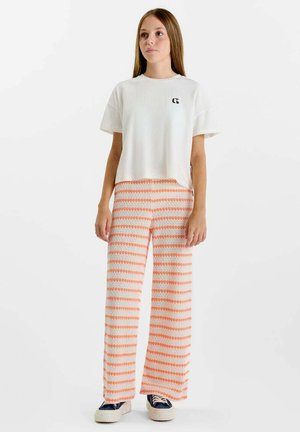 Adolescente debout portant une chemise blanche à manches courtes avec un petit logo et un pantalon à jambes larges à rayures horizontales orange et blanches.