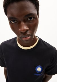 T-shirt nera con colletto giallo a contrasto. Presenta un logo a forma di sole blu sul lato sinistro del petto con il testo: "ARMEDANGELS SINCE 2007." Design minimalista.