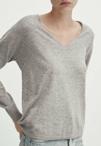 Pull en V gris en maille douce, avec des manches longues et une coupe décontractée, porté avec un jean bleu clair.