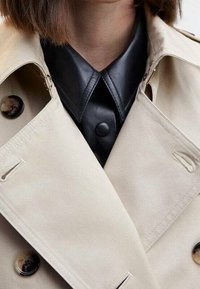 Beżowy płaszcz trench z podwójnym zapięciem, z dużymi czarnymi guzikami i warstwową czarną skórzaną koszulą pod spodem.