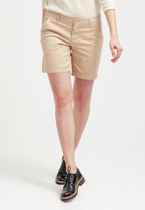 Personne portant un short beige jusqu'aux genoux, un pull de couleur claire et des chaussures en cuir noires à lacets, debout devant un fond uni.