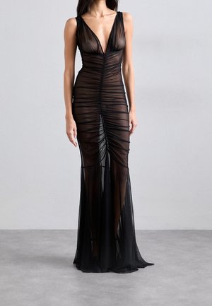 Vrouw die een lange zwarte doorschijnende jurk draagt met een diepe V-hals en geplooide details, staand tegen een effen grijze achtergrond.