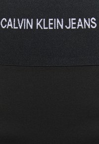 Černý textilní pásek s vyšívaným nápisem "CALVIN KLEIN JEANS" v bílé barvě. Hladký povrch, obdélníkový tvar, minimalistický design.
