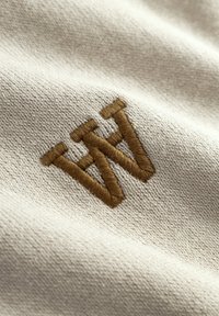 Tissu beige orné d'une lettre "W" brodée en or foncé. Le tissu présente une texture subtile, mettant en valeur la broderie détaillée du logo.