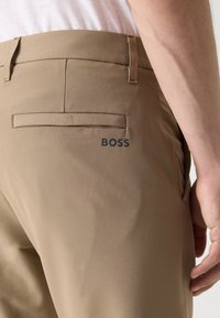 Pantaloni bej croiți dintr-un material neted, cu un buzunar posterior dotat cu clapetă și un logo negru "BOSS" lângă buzunar.