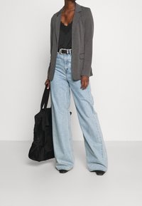 Blazer gris sobre camisón de encaje negro, jeans anchos de color azul claro y bolsa de mano negra. Características incluyen zapatos negros de punta y cinturón plateado como acento.