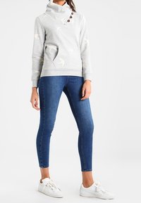 Sudadera gris con cuello de embudo, con patrones de pájaros en crema y botones de madera. Combinada con jeans ajustados azules y zapatillas blancas.