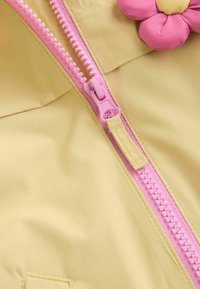 Tela amarilla con una textura suave que presenta una cremallera rosa. Un adorno de flor rosa está colocado cerca de la cremallera.