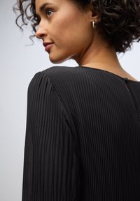Schwarzes, gefältetes Kleid mit Rundhalsausschnitt und Knopfdetails auf der Rückseite, aus strukturiertem Stoff und mit langen Ärmeln.