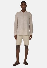 Camicia di lino beige con colletto rilassato, abbinata a pantaloni corti beige chiaro. Scarpe tipo mocassino marroni completano l'outfit.