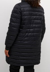 Cappotto imbottito nero con texture trapuntata, collo alto e maniche lunghe. Presenta polsini a coste e un taglio dritto che arriva a metà coscia.