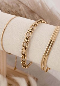 Három arany karkötő, egy texturált krémszínű tartón kiállítva; egy láncszemes, egy lapos, több szálas és egy vékony kígyólánc.