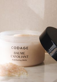 Transparenter Behälter mit hellrosa Peelingbalsam und schwarzem Deckel. Text lautet "CODAGE BAUME EXFOLIANT Exfoliating Balm." Textur sichtbar.