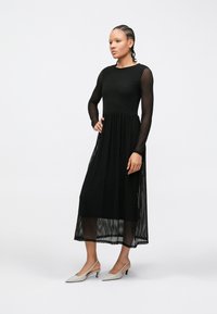 Sieviete ar pinumiem matos, tērpusies melnā vidēja garuma kleitā ar teksturētu audumu, caurspīdīgām garām piedurknēm un sāniem, valkājot baltas zemu papēžu slingbeka kurpes.