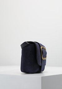 Navy modrý semišový pytel s obdélníkovým tvarem, obohacený černým popruhem z umělé kůže a zlatým sponou na klopě.