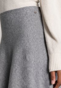 Prenda de lana con una sección superior color crema y una parte inferior de un gris suave. Un detalle notable es un pequeño emblema metálico en la costura.