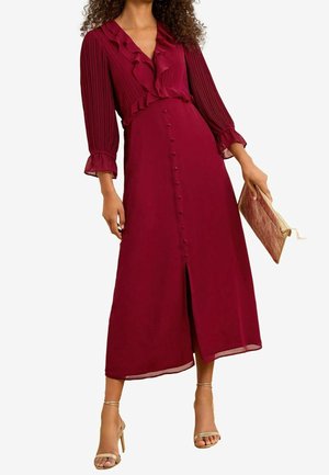Femme portant une longue robe bordeaux avec un col à volants, boutonnée devant, manches longues transparentes, tenant une pochette dorée brillante et des talons hauts dorés.