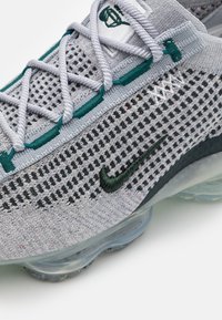 Grå stickad träningssko med mesh-design, teal accenter, svart Nike swoosh och genomskinlig luftkuddessula. Snörena är ljusgrå.