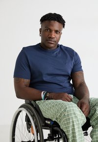 Tengerész stílusú crew-neck póló, pamut anyagból. A modell, aki kerekesszékben ül, zöld kockás pizsamát visel, és több karkötő díszíti a csuklóját, egy egyszínű háttér előtt ül.