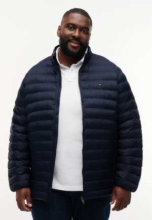 Mand smiler iført en mørkeblå quiltet jakke over en hvid poloshirt, stående foran en ensfarvet hvid baggrund.