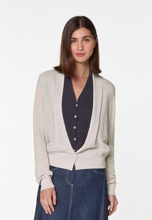 Cardigan beige clair avec une texture douce, à l'avant ouvert et aux manches longues. Porté sur une chemise noire à boutons et associé à une jupe en denim.