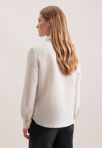 Frau mit langen, welligen Haaren, die ein weißes, geripptes Langarmshirt und schwarze Hosen trägt, von hinten vor einem schlichten Hintergrund gezeigt.