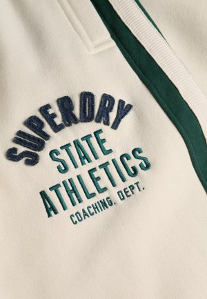 Tejido crema con texto bordado: "SUPERDRY STATE ATHLETICS COACHING DEPT." en azul marino y azul verdoso, con acentos de ribete verde.